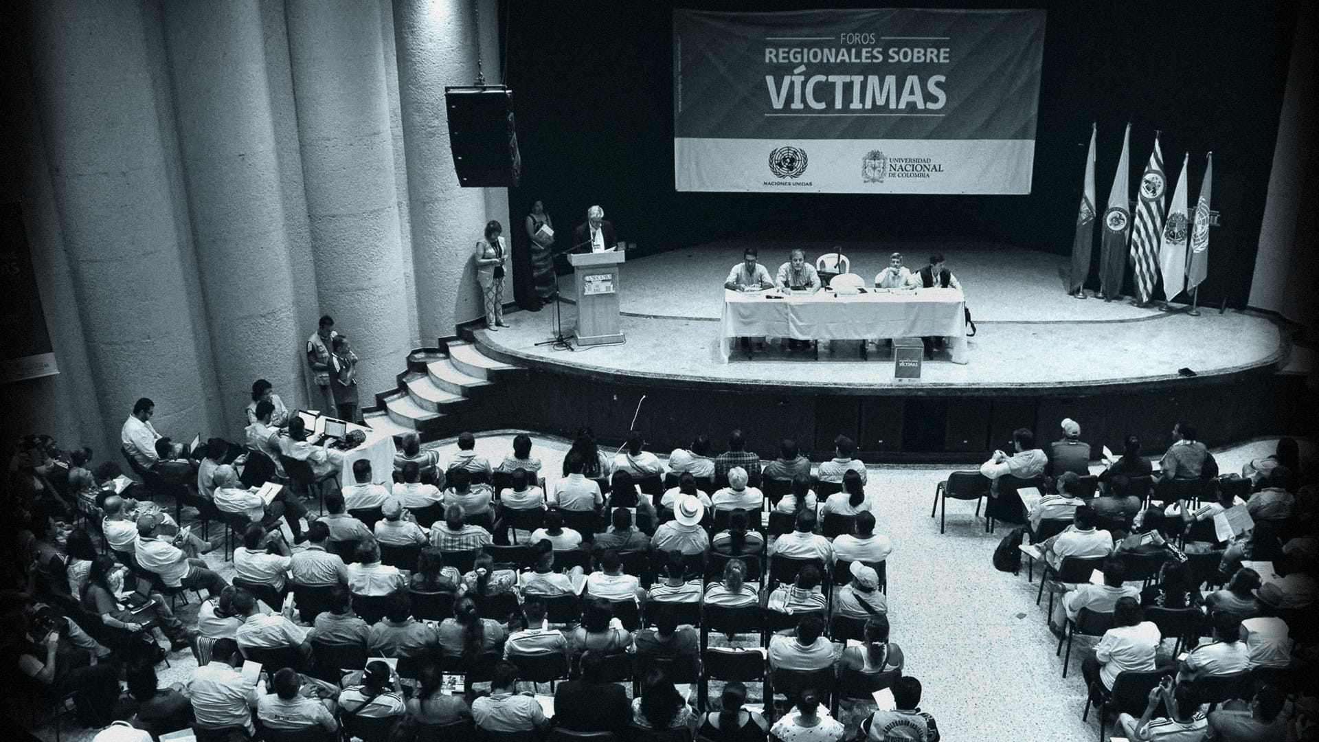 Escenario con personas que presentan a otras que están sentadas en sillas, al fondo una pancarta que dice: Foros Regionales sobre víctimas 