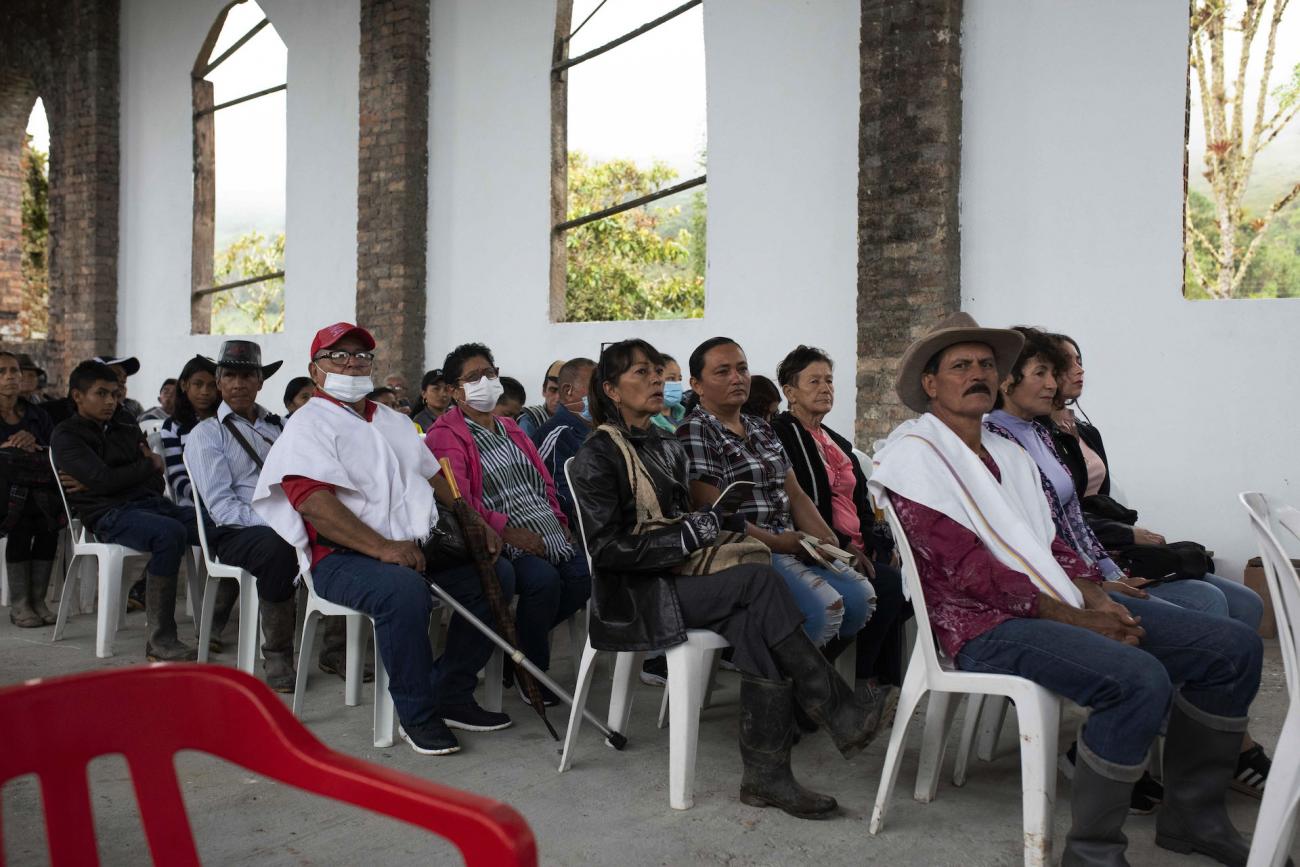 Grupo de personas y campesinos con poncho y sombrero, sentados en salón sin ventanas, afuera paisaje nublado y árboles