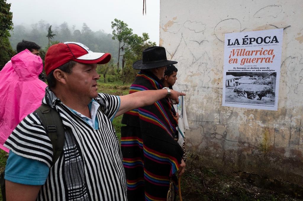 Personas con poncho, sombrero e impermeable observan un afiche pegado al muro. Texto: “La época. Villarrica en Guerra. cielo nublado