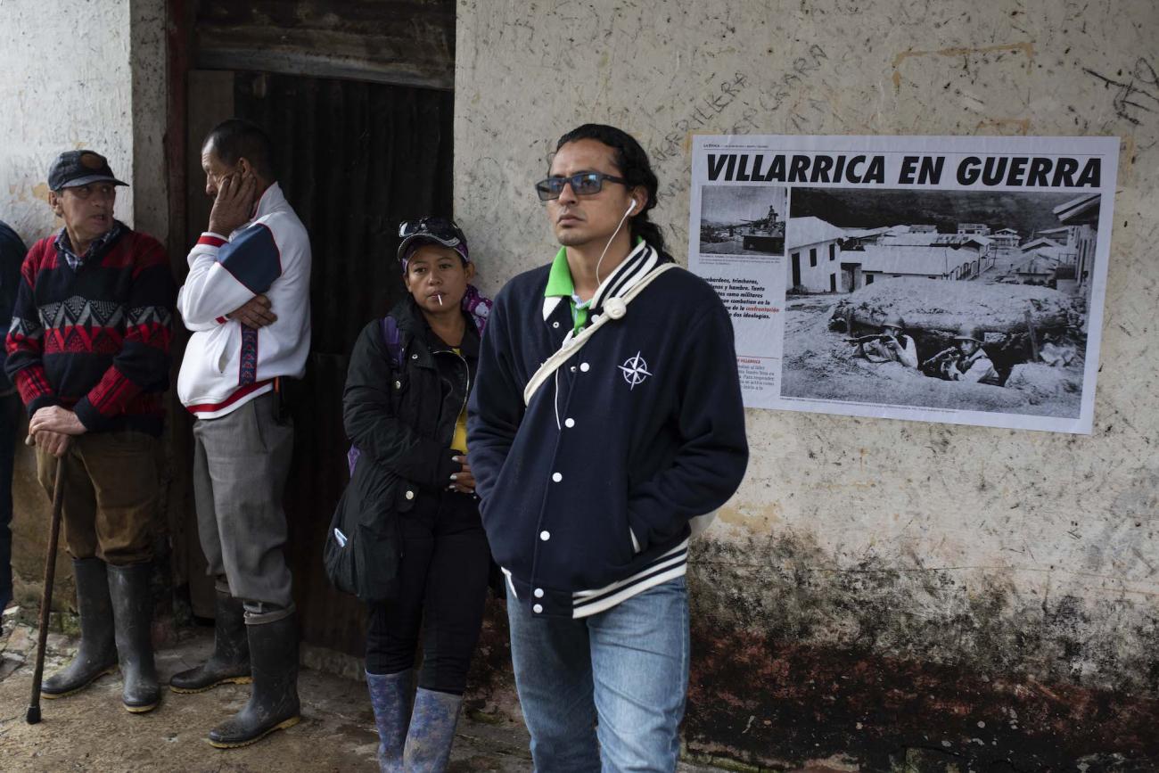 2 Personas conversan, 2 miran al horizonte. Sobre el muro hay un afiche en una edificación abandonada. Texto: Villarrica en guerra