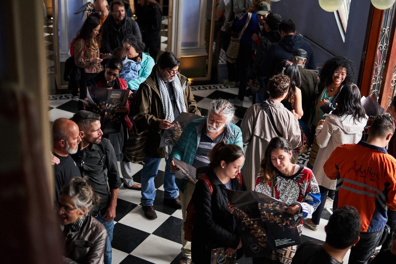 Asistentes al evento esperan en fila acceso mientras leen el programa de mano