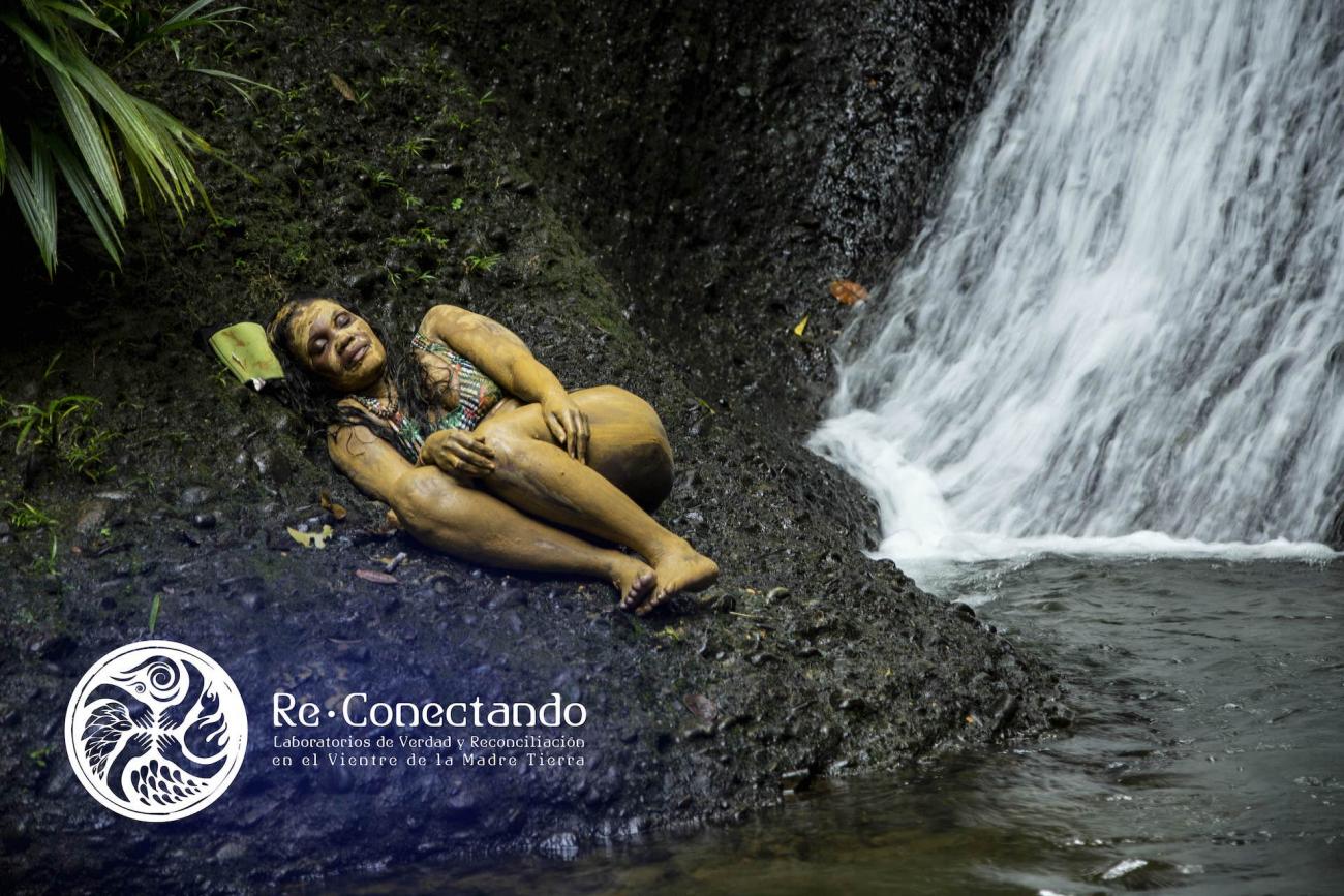 Mujer afro cubierta con barro reposa sobre una piedra al lado de una cascada