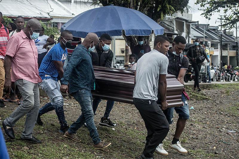 5 hombres afro cargan un ataúd con otros hombres alrededor, al fondo hay un sombrilla grande y un árbol