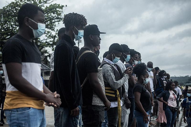 Hombres y mujeres afro están de pie unos junto a otros mirando al horizonte, algunos llevan tapabocas