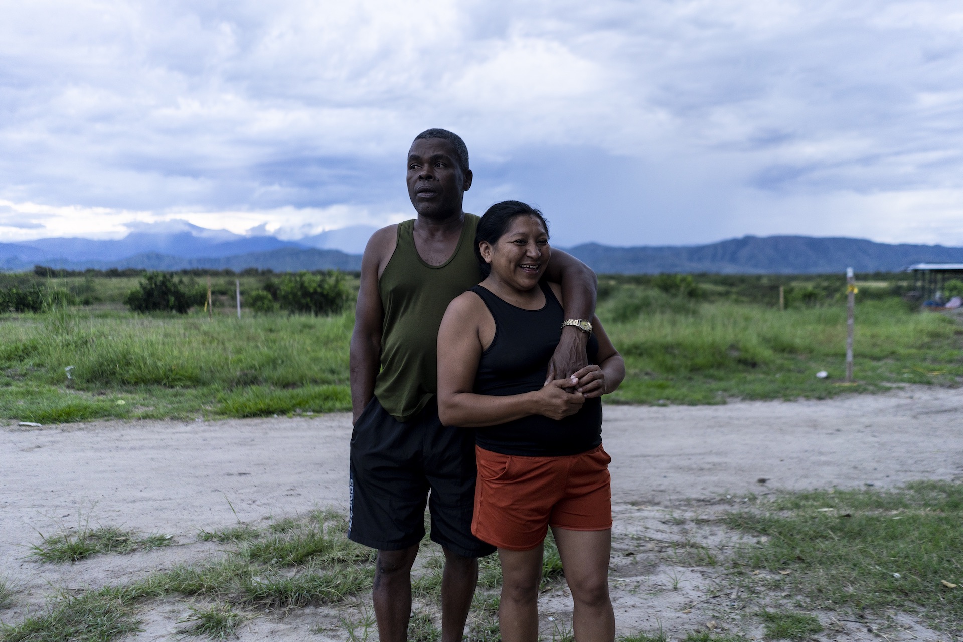 En paisaje de pradera y montañas, un hombre en camiseta sin mangas y pantaloneta abraza a mujer sonriente