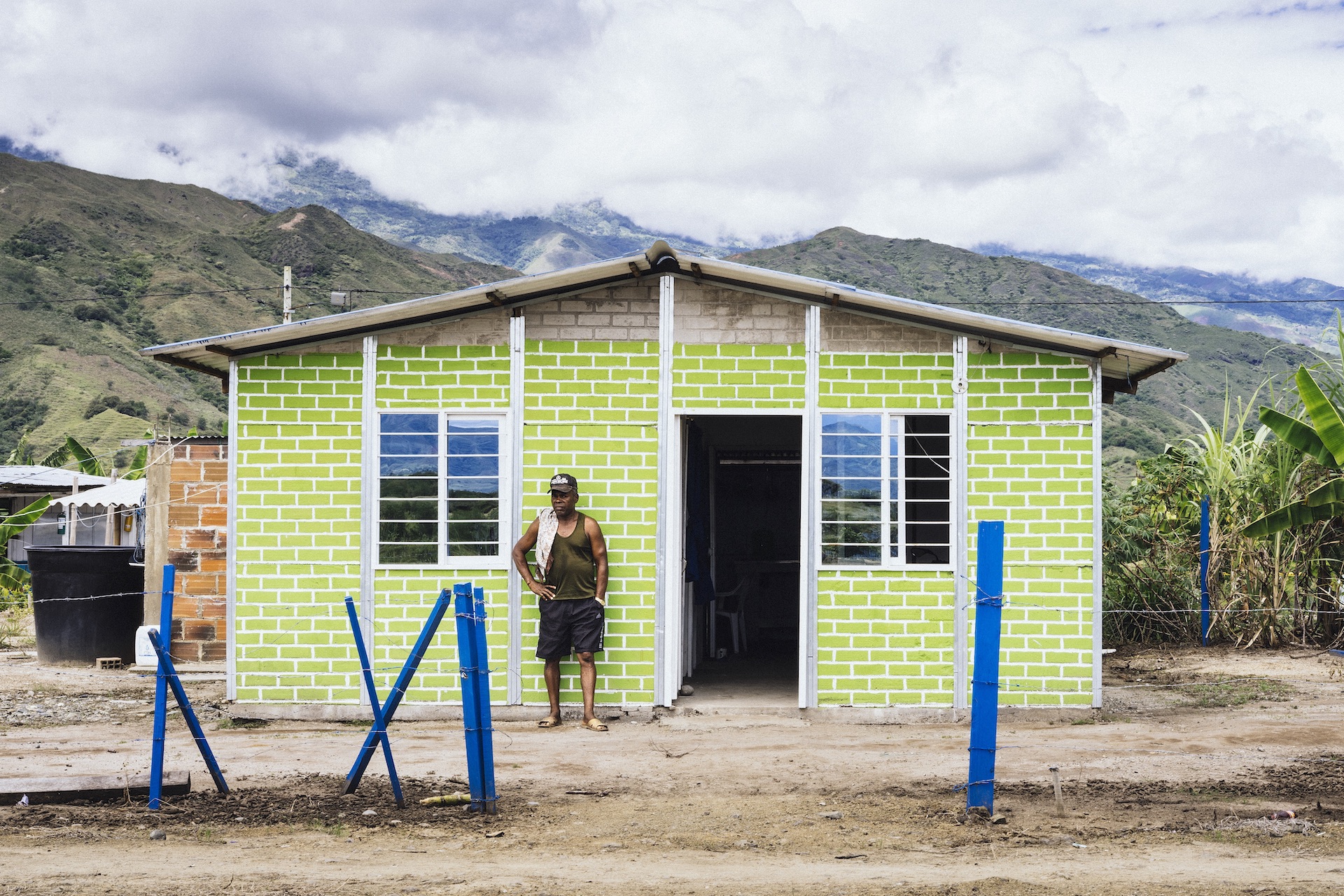 Hombre en pantaloneta, camiseta sin mangas y cachucha de pie frente a una casa rural, al fondo montañas y nubes