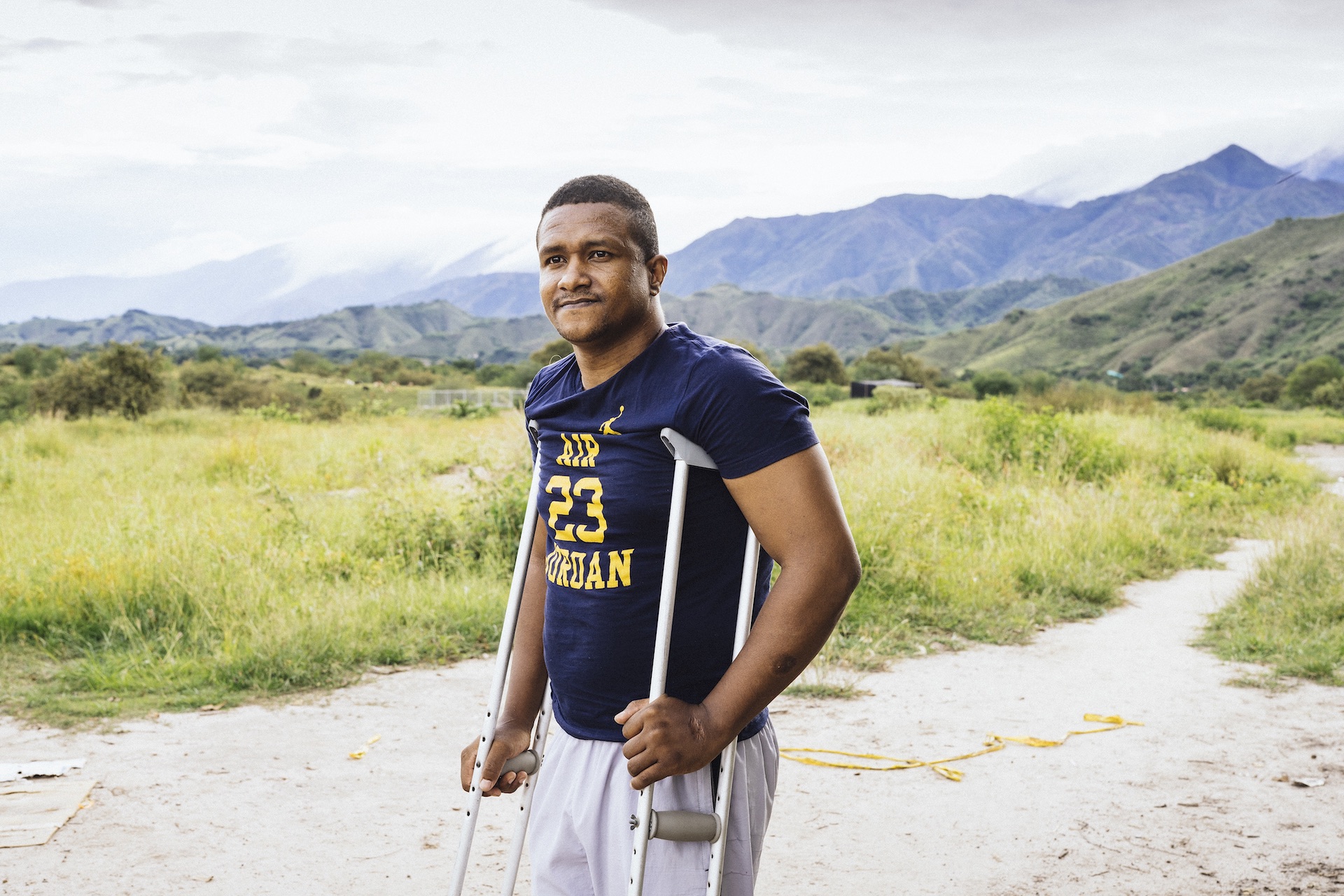 Hombre en muletas mirando al horizonte, al fondo un camino de tierra y montañas